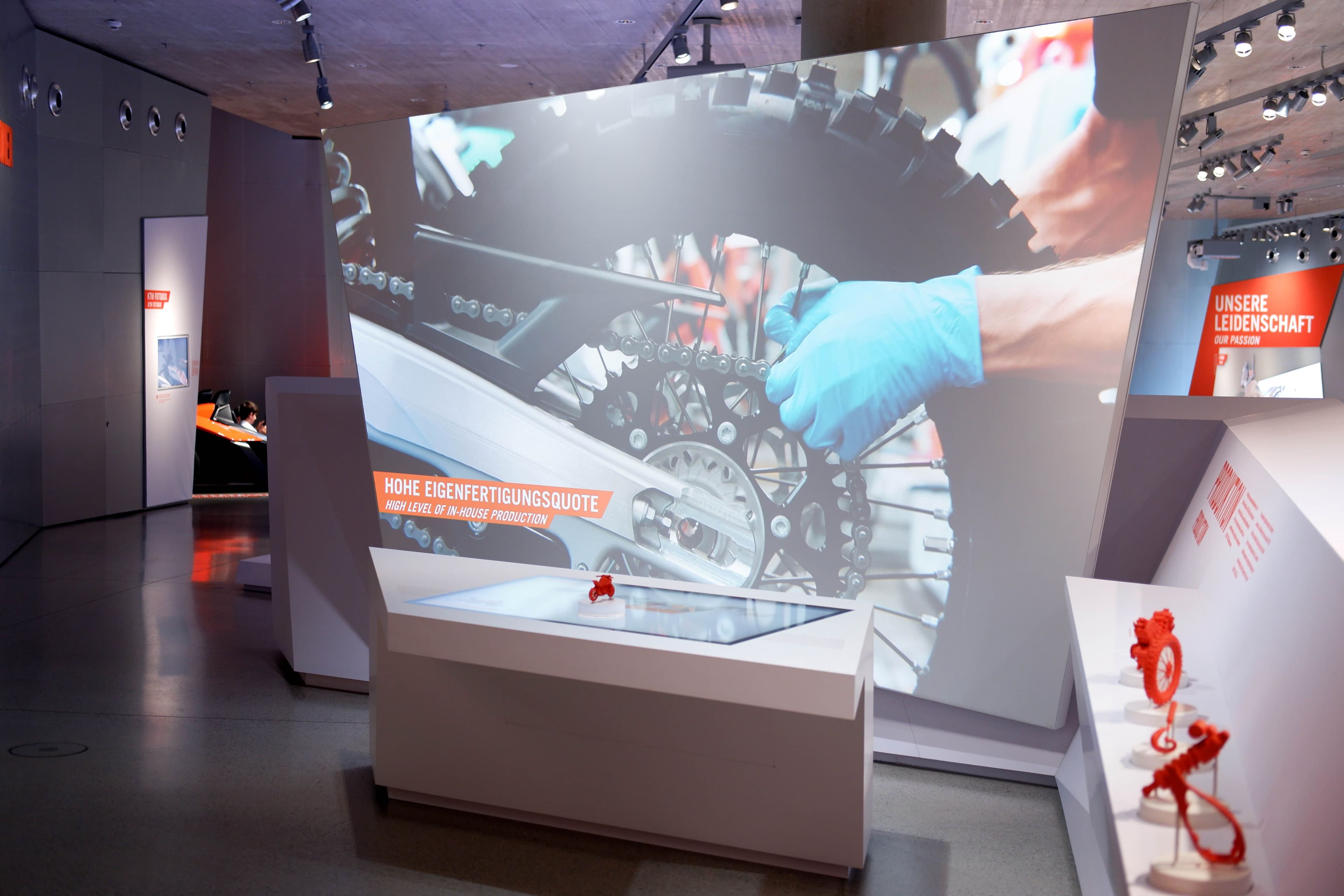 Übersicht des Exponats: Tisch mit aufgelegtem Token, im Hintergrund ein Video von der Montage eines Motorrades